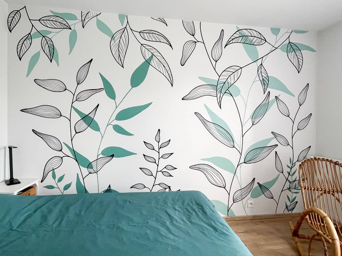 Fresque murale végétale chambre peinte sur mesure : feuillages botaniques graphiques en noir et vert céladon sur fond blanc. Un décor mural apaisant et contemporain, en parfaite harmonie avec la literie et le mobilier rotin.