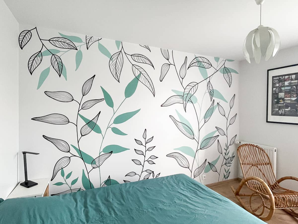 Fresque murale végétale chambre peinte sur mesure : feuillages botaniques graphiques en noir et vert céladon sur fond blanc. Un décor mural apaisant et contemporain, en parfaite harmonie avec la literie et le mobilier rotin.