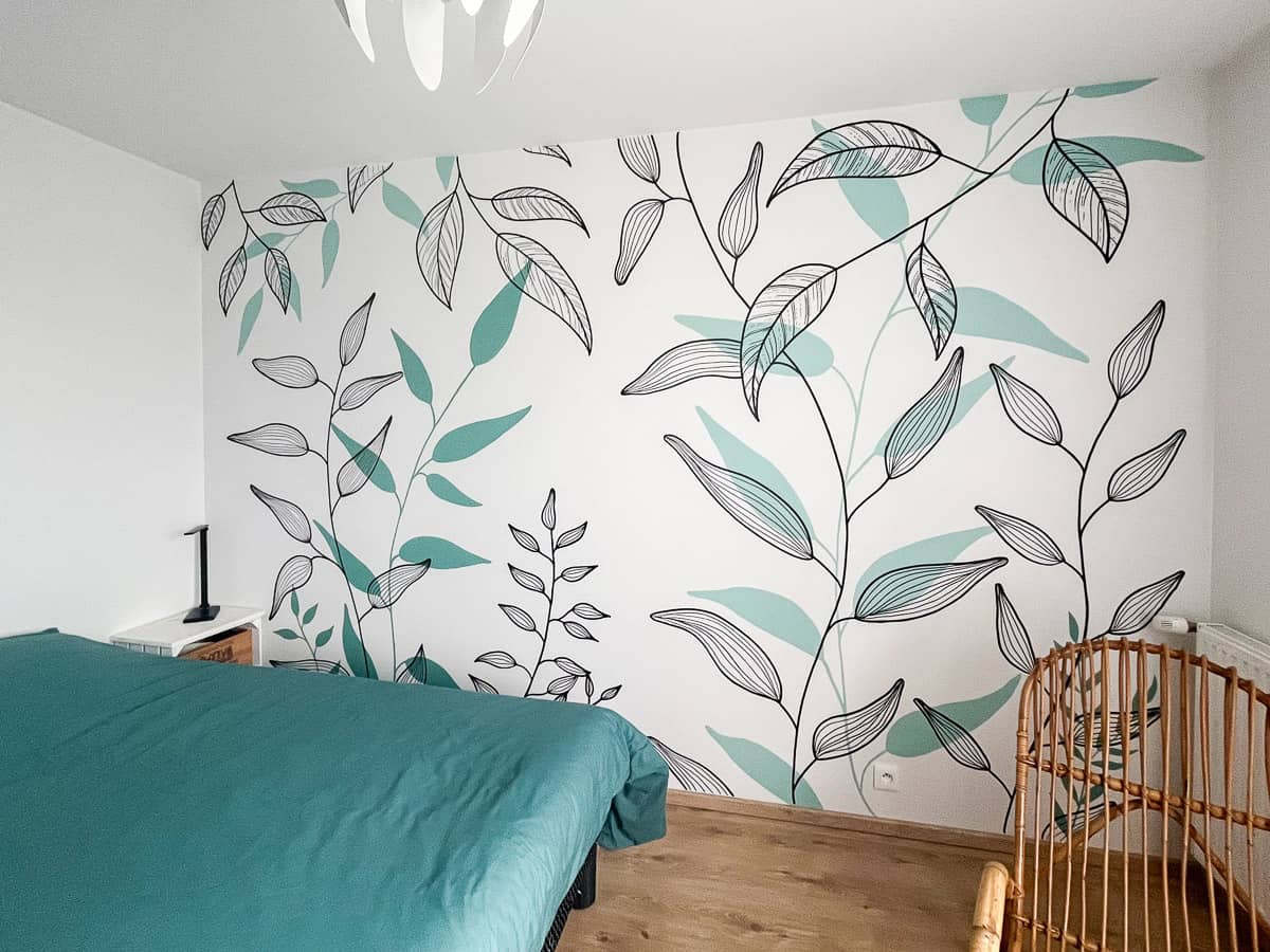 Fresque murale végétale chambre peinte sur mesure : feuillages botaniques graphiques en noir et vert céladon sur fond blanc. Un décor mural apaisant et contemporain, en parfaite harmonie avec la literie et le mobilier rotin.