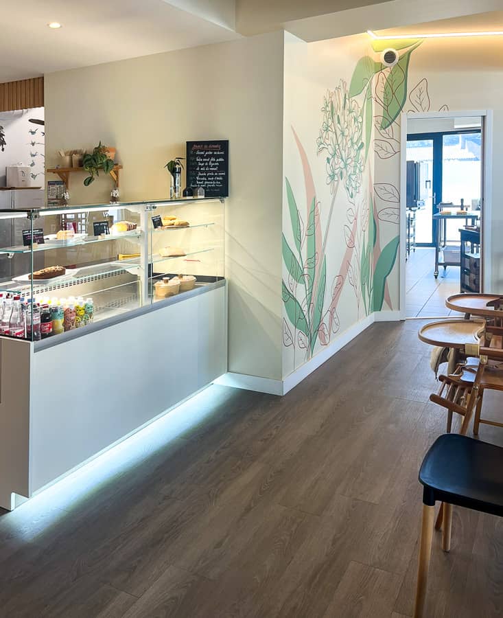 Fresque murale restaurant peinte sur mesure : agapanthe et feuillages en rose poudré et vert sauge sur fond blanc crème. Une décoration murale végétale qui crée une identité visuelle forte et une ambiance chaleureuse pour vos clients. Vue de l'entrée