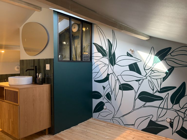 Salle de bain moderne avec un meuble vasque en bois, un miroir asymétrique et un mur décoré d'un motif de feuilles d'eucalyptus stylisées vert foncé.